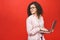 Portrait of an excited curly young girl holding laptop computer isolated over red background