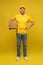 Portrait of excited delivery man in yellow uniform holding paper box isolated over yellow background.