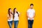 Portrait of excited girls mocking at guy standing shy wearing white t-shirt denim jeans isolated over yellow background