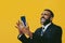 portrait of expressive angry excited bearded man in suit and tie with smartphone video call hand up yellow background