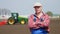 Portrait of farmer or agronomist, in red plaid shirt, against background of working tractor, cultivator, agricultural