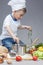 Portrait of Fascinated Caucasian Little Boy Working With Dill In Cooking Hat. Surrounded by Fresh Fruits