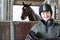 Portrait Of Female Owner Holding Saddle In Stable With Horse