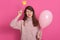 Portrait of flirty attractive winking dark haired woman with pink balloon, dresses warm rose sweater, posing against rosy studio
