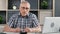 Portrait of focused 70s grandfather posing at workplace with laptop during paperwork at home office