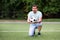 Portrait of football coach or gym teacher standing on one knee holding and giving soccer ball to camera and talking on green field