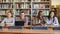 Portrait of four multi-ethnic students sitting at long desk in big spacious library with piles of books looking at