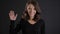 Portrait of fragile young brunette woman watching happily and waving hand into camera on black background.