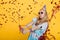 Portrait of funny blond woman in birthday hat and red confetti on yellow background. Celebration and party.