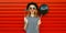 Portrait of funny cheerful young woman having fun holding black balloon, showing mustache on stick on red background