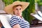 Portrait of funny grimace childish young adult girl in hat and dress sitting and resting on cozy sunbed, showing tougue out