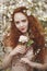 Portrait of Gentle woman with long red hair with a rose in a blooming garden. Red-haired girl with pale skin and blue