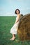 Portrait of a girl with short black hair in a bright white dress amongst haystacks in a green field.