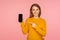 Portrait of good looking positive ginger girl in sweater pointing at cellphone and smiling at camera, recommending mobile