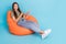 Portrait of gorgeous girl with straight hair dressed striped t-shirt sit on pouf directing empty space isolated on blue