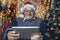 Portrait of a grandfather dressed in sweater posing in cosy room with christmas tree holding a tablet