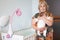 Portrait of grandma carrying a newborn infant in her arms, standing near white crib in bedroom, looking at camera