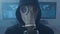 Portrait of Hacker in gas mask and hood in smoke cyber security center filled with display screens. World of