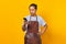 Portrait of handsome asian man wearing apron looking unhappy reading incoming messages on mobile on yellow background