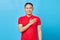 Portrait of handsome asian young man in red shirt holding hands on chest to thank someone and express gratitude isolated on blue