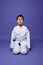 Portrait of a handsome child boy wearing kimono and sitting in aikido stance against purple background with copy space for text.