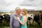 Portrait of Handsome Senior Farmer and His Young Blonde Wife Hugging in Front of Their Black and White Cows Outdoors