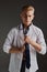 Portrait of handsome young man posing in white shirt, tying tie isolated over grey background. Classical male fashion