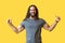 Portrait of happy bearded young man with long curly hair in grey tshirt standing with thumbs up and looking at camera with toothy