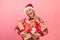 Portrait of happy blonde woman in cozy sweater and Santa hat holding heap of Christmas presents on pink background