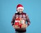 Portrait of happy Caucasian guy in Xmas sweater and Santa hat holding pile of holiday gifts on blue studio background