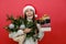 Portrait of happy cute young woman in santa hat looking hold bouquet of spruce branches and Christmas presents, posing isolated