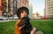Portrait happy girl in hat sits with dog on lawn against cityscape background, hugs puppy and looks into camera with smile.Photo