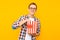 Portrait of a happy man in transparent glasses with a bucket of popcorn, keen on watching a movie, on a yellow background
