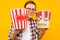 Portrait of a happy man in transparent glasses with a bucket of popcorn, keen on watching a movie, on a yellow background