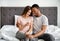 Portrait of happy multiracial couple holding positive pregnancy test, sitting on bed, hugging and enjoying good news
