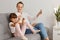 Portrait of happy optimistic woman wearing white shirt and jeans sitting on sofa with her daughter and looking at child`s phone