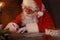 Portrait of happy Santa Claus sitting at his room at home near Christmas tree and answering Christmas letters