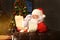 Portrait of happy Santa Claus sitting at his room at home near Christmas tree and reading Christmas letter or wish list.