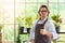 Portrait of happy senior man gardening wearing glasses and denim apron. Showing thumbs up, smiling and standing with plant shelf