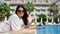 Portrait of happy travel woman posing on deck chair waving hand at blue water palm hotel background