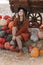 Portrait of happy woman sitting on straw near wagon with orange pumpkin on farmers market in brown sweater, dress and hat. Cozy