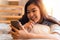 Portrait of happy young Asian girl in casual clothing lying down on bed while making a video call with smartphone and