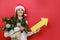 Portrait of happy young female in Santa Claus hat holding bouquet of spruce branches and paper yellow arrow, posing on red studio