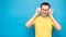 Portrait of happy young stylish man in yellow t-shirt, glasses and headphones expressing positivity isolated on blue background,