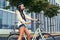 Portrait of a hippie female wearing blouse and shorts in a headband, sitting on a city bike against a skyscraper