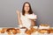 Portrait of hopeful cute woman with brown hair wearing white T-shirt sitting at table  over gray background., holding cake