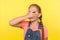 Portrait of inquisitive little girl with braid in denim overalls covering face with hand and looking through fingers