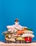 Portrait of irritated, offended little princess, girl sitting on huge, big pile of pillows over blue studio background