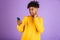 Portrait of joyful african american man smiling and holding smartphone, wearing bluetooth earpod, isolated over violet background