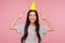 Portrait of joyful funny asian girl pointing at party cone on head and looking at camera with toothy smile